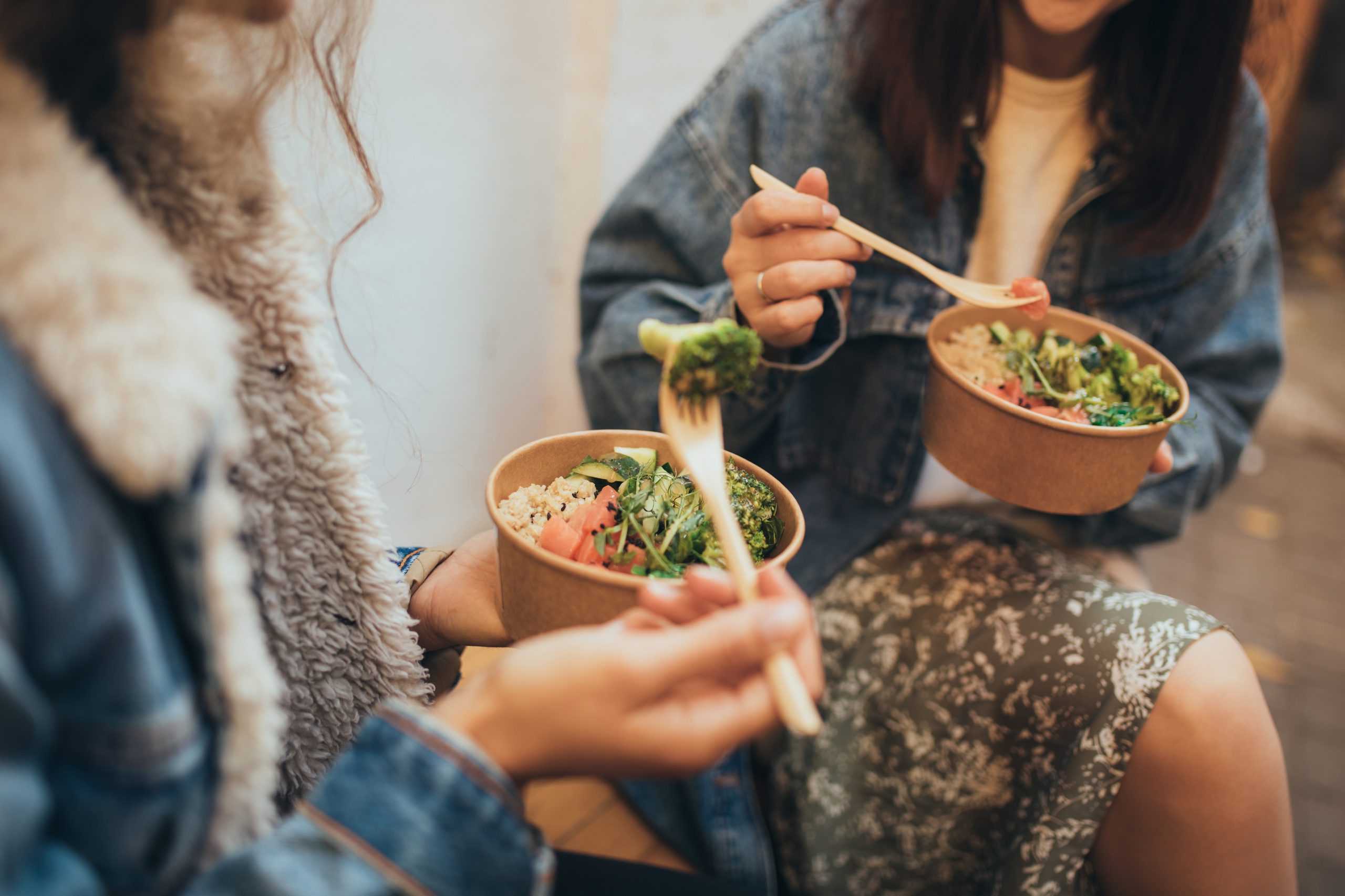 Two millennial women female friends sitting outdoors eating takeaway food, laughing and having fun. Food delivery and takeout.